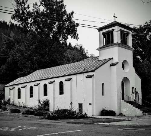 A white church with a bell tower and cross, surrounded by trees, captured in black and white.