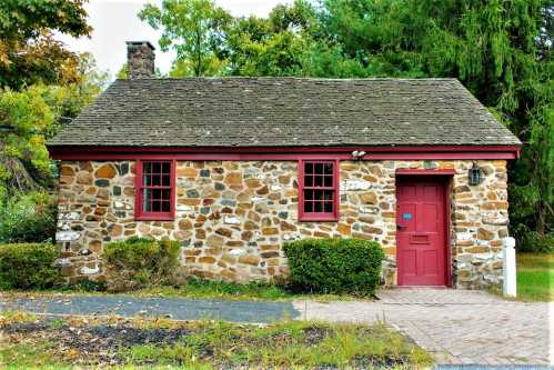 A small stone cottage with a red door and windows, surrounded by greenery and a paved pathway.