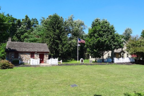 Historic buildings with a stone cabin and a house, surrounded by trees and a grassy area, featuring an American flag.