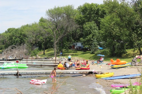 A beach scene with people relaxing on colorful inflatables, a dock, and trees in the background on a sunny day.