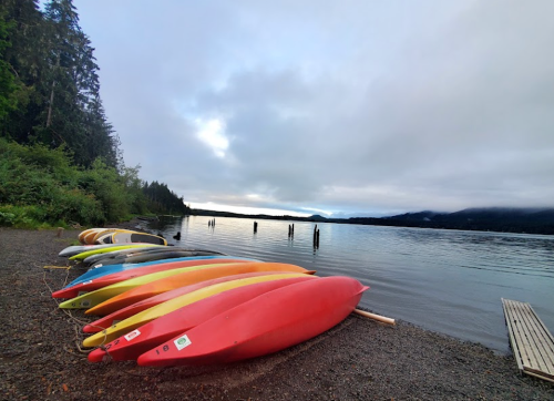 Colorful kayaks lined up on a gravel shore beside a calm lake, surrounded by trees and a cloudy sky.
