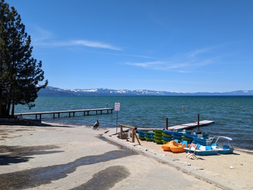 A serene lake view with a wooden pier, colorful paddle boats, and snow-capped mountains in the background under a clear blue sky.