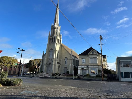 A tall church with a spire next to a large house, set against a clear blue sky and street view.
