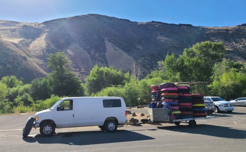 A white van towing a trailer stacked with colorful inflatable tubes, parked near a green landscape and mountains.
