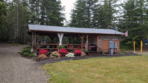 A rustic outdoor pavilion surrounded by greenery and colorful flowers, with picnic tables and an American flag.