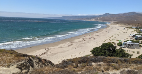 A scenic view of a sandy beach with gentle waves, surrounded by hills and a few scattered tents and buildings.