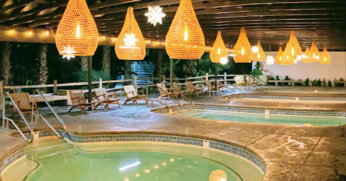 A serene pool area with warm lighting, featuring wicker pendant lamps and lounge chairs under a wooden pergola.