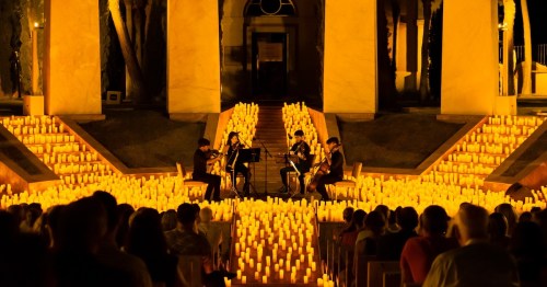 A string quartet performs surrounded by glowing candles, with an audience seated in a beautifully lit outdoor setting.