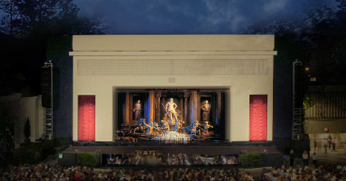 A grand outdoor theater stage with a large backdrop and an audience seated in front, illuminated at night.