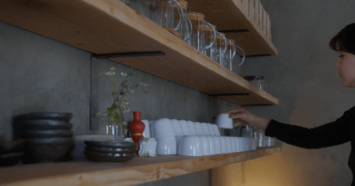 A person arranges white cups on a wooden shelf, surrounded by glassware and decorative items.