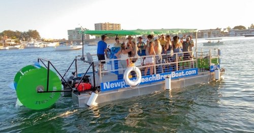 A party boat with people enjoying themselves on the water, surrounded by boats and a sunny shoreline.
