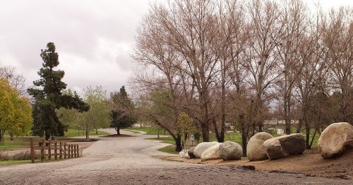 A gravel path leads through a park with bare trees and large rocks, under a cloudy sky.