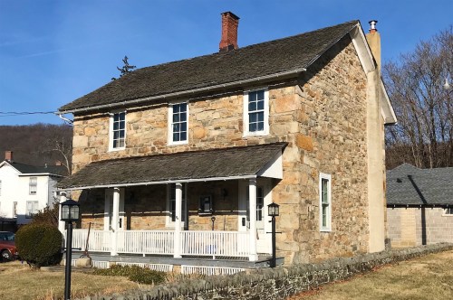 A historic stone house with a porch, surrounded by grass and trees, under a clear blue sky.