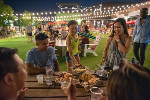 A lively outdoor gathering with friends enjoying food and drinks under string lights at night.