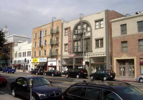 A street view featuring a mix of buildings, shops, and parked cars under a cloudy sky.