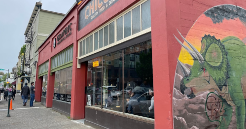 A colorful mural of a dinosaur on a red building, with people walking by on a cloudy day.
