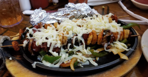 A sizzling platter of skewered meat and vegetables topped with melted cheese, served on a wooden board.