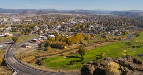 Aerial view of a small town surrounded by mountains, featuring green fields and autumn-colored trees.