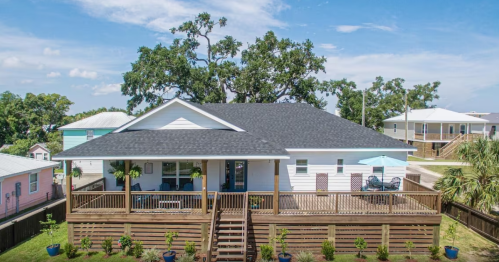 A modern house with a large front porch, surrounded by greenery and blue skies, featuring a deck and patio furniture.