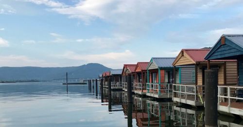 Colorful waterfront cabins line a calm lake, reflecting in the water with mountains in the background.