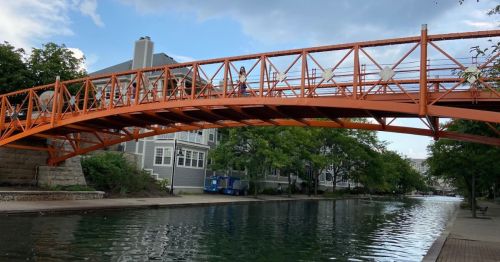 An orange pedestrian bridge spans a calm canal, surrounded by greenery and buildings under a partly cloudy sky.