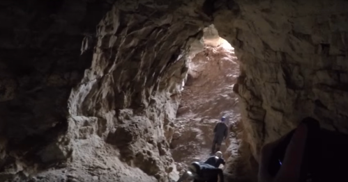 A dimly lit cave with rocky walls, showing two people climbing towards a bright opening.