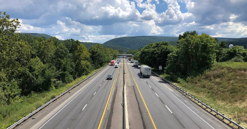 A busy highway with vehicles traveling in both directions, surrounded by green trees and rolling hills under a cloudy sky.
