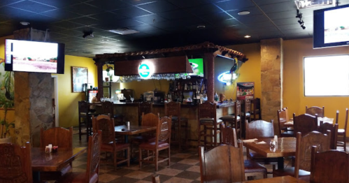 Interior of a restaurant featuring wooden tables, a bar with drinks, and TVs on the walls. Bright yellow walls.