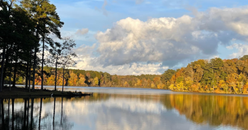 A serene lake surrounded by trees with autumn foliage, reflecting clouds and blue sky in the water.