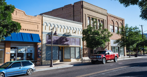 A street view of a small town featuring brick buildings, trees, and parked cars under a clear blue sky.