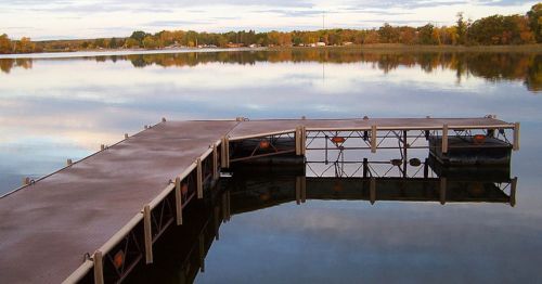 A serene lake scene featuring a wooden dock extending into calm waters, reflecting trees and a cloudy sky.