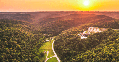 Aerial view of lush green hills at sunset, with a winding road and a building nestled in the valley.