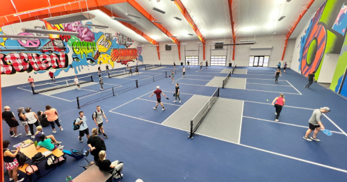 A spacious indoor sports facility with colorful murals, featuring multiple tennis courts and players engaged in games.
