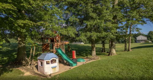 A colorful playground with a slide, playhouse, and climbing structure, surrounded by trees and green grass.