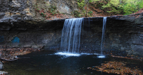 A serene waterfall cascading over rocks into a dark pool, surrounded by lush greenery and autumn leaves.