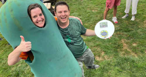 A person in a pickle costume gives a thumbs up next to a smiling man holding a frisbee at an outdoor event.