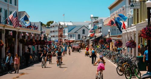 A bustling street scene with people biking, shops, and flags, surrounded by colorful flower baskets on a sunny day.