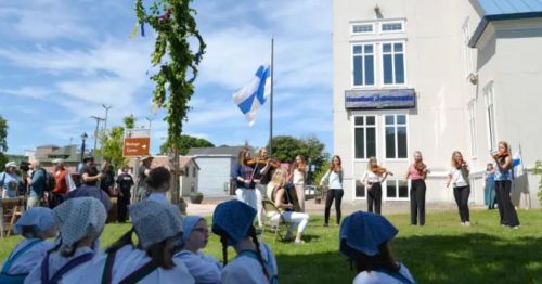 A group of musicians perform with violins outdoors, while an audience watches in a sunny setting with a Finnish flag.
