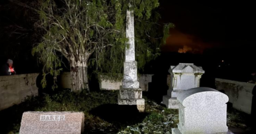 A dimly lit cemetery at night featuring several gravestones and a tall monument surrounded by trees.
