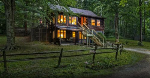 A cozy wooden cabin surrounded by trees, featuring large windows and a welcoming porch, set in a serene forest.