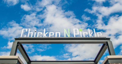 Sign for "Chicken N Pickle" against a blue sky with clouds.