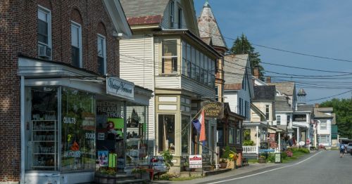 Charming small-town street lined with historic buildings, shops, and colorful flower displays under a clear blue sky.