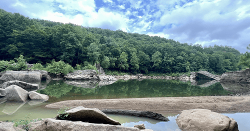 A serene river scene with lush green trees, rocky banks, and a cloudy blue sky reflecting on the calm water.