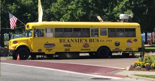 A bright yellow food bus named "Beanie's Bus" with a menu featuring hot dogs, chicken nuggets, and fries.