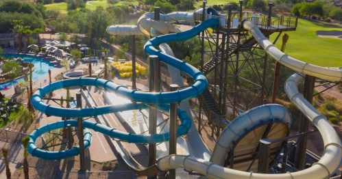 Aerial view of a water slide complex with blue slides, surrounded by greenery and a pool area.