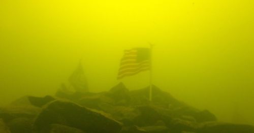 An American flag and another flag submerged underwater, surrounded by rocks in a murky yellow environment.