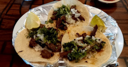 Three tacos filled with seasoned meat, chopped onions, and cilantro, served with lime wedges on a silver platter.