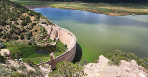 Aerial view of a curved dam surrounded by green water and lush landscape under a clear blue sky.