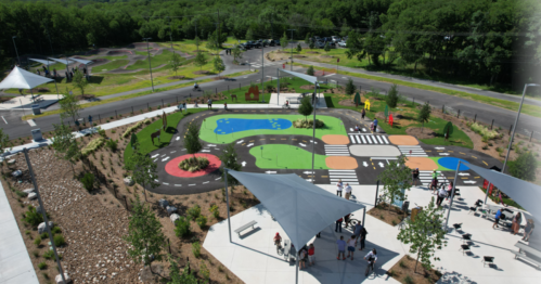 Aerial view of a colorful playground with pathways, play areas, and green spaces, surrounded by trees and a road.