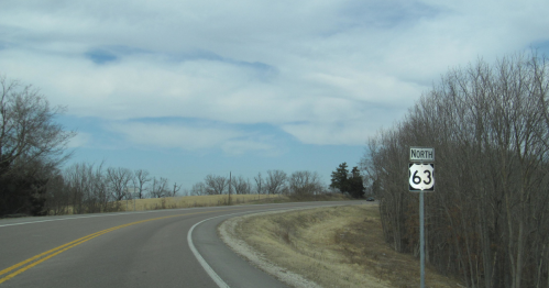 A winding road with a sign indicating "North 63" and bare trees against a cloudy sky.